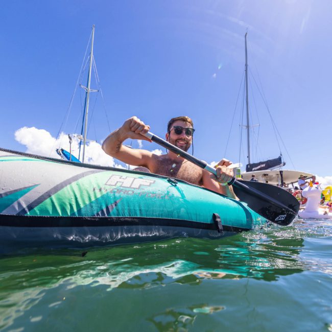 A person is kayaking in clear blue water, surrounded by sailboats and other kayakers under a sunny sky, enjoying the vibrant atmosphere of a Sydney boat party hire.
