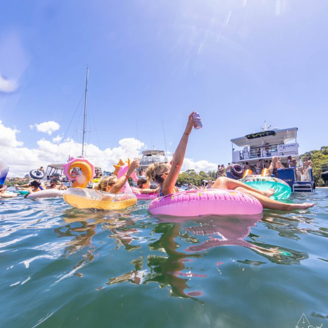 People in inflatable pool floats are enjoying a sunny day in the water near yachts, with one person holding up a drink, all against the backdrop of an exciting catamaran party Sydney.