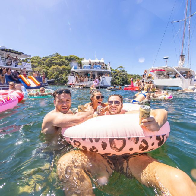 People in swimsuits are enjoying a sunny day in the water, surrounded by boats and floating on inflatable devices. Some hold drinks and smile at the camera, creating the perfect scene for a private yacht charter on Sydney Harbour.