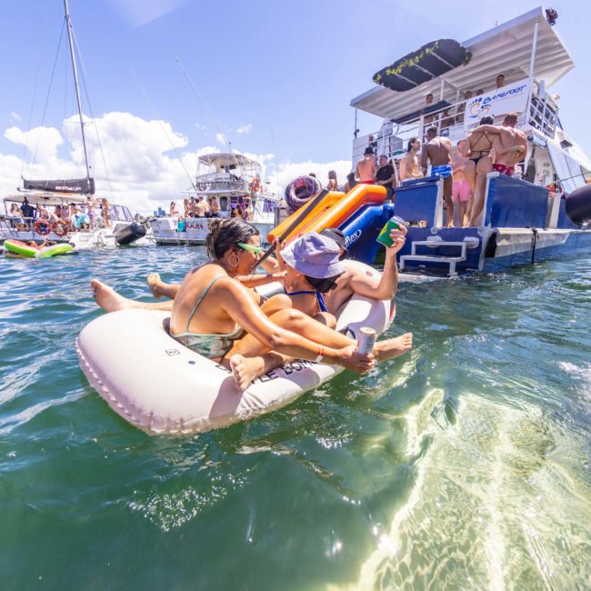 Two people relax on an inflatable float in a sunny, crowded lake with multiple boats and swimmers in the background, creating an atmosphere reminiscent of a luxury yacht hire Sydney.