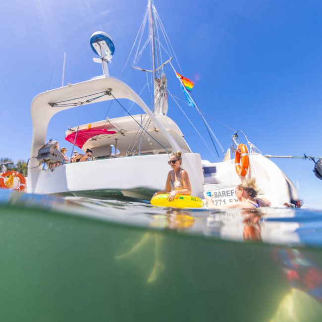 People onboard and swimming near a white sailboat on a sunny day, with part of the image showing an underwater view. A rainbow flag is visible on the boat, indicating a lively catamaran party in Sydney.