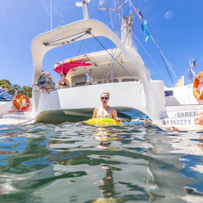 People enjoying a sunny day on a catamaran; one person is floating on a yellow inflatable in the water, while others are sitting on the boat, making the most of their Catamaran party Sydney.