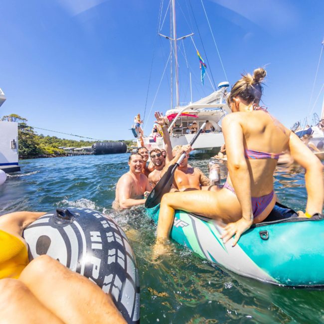 A group of people enjoying a sunny day in the water with inflatable rafts, boats, and yachts in the background. Some individuals are standing and sitting on the rafts, engaging in conversation and laughter during their Sydney boat party hire.