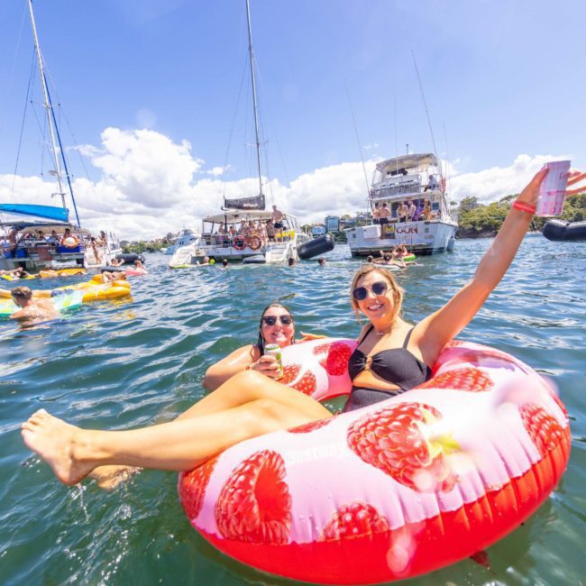 People on floating inflatables in the water, with boats in the background and a sunny sky. One person holds a drink and waves. Enjoy a perfect day with a private yacht charter on Sydney Harbour or book a DJ boat hire for an unforgettable Sydney boat party experience.