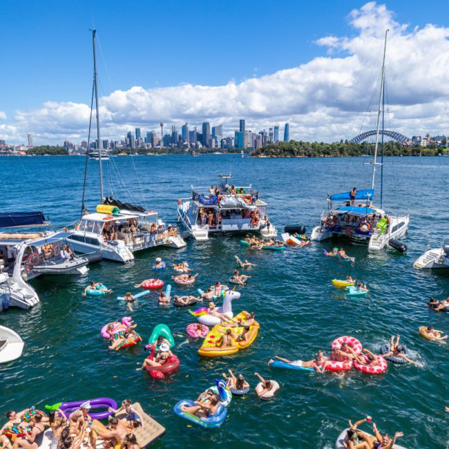 Dozens of people in colorful floaties enjoy swimming near anchored boats on a sunny day. The background shows a city skyline and the famous Sydney Harbour Bridge, with private yacht charters adding to the fun atmosphere.