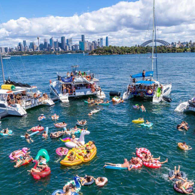 People are enjoying a sunny day on a body of water with inflatables and boats, with a skyline and bridge visible in the background, perfect for Corporate boat events Sydney or a fun Catamaran party Sydney.