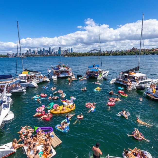 People enjoying a sunny day on a large body of water with several boats and floating devices. In the background, there is a city skyline set against a blue sky with scattered clouds, perfect for those considering luxury yacht hire Sydney.
