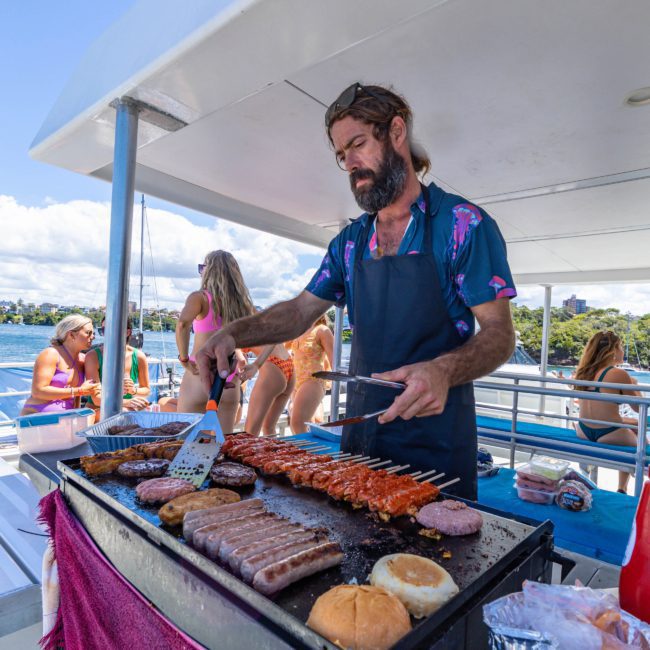 A man is grilling various meats on a barbecue aboard a boat in Sydney. People in swimsuits can be seen in the background enjoying the sunny weather, creating the perfect ambiance for your next corporate boat event or catamaran party.