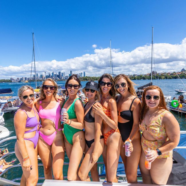 Seven women in swimsuits smile and pose together on a private yacht charter in Sydney Harbour, holding drinks with a city skyline and other boats in the background under a clear blue sky.