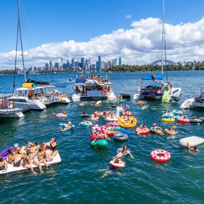 People on boats and inflatables enjoy a sunny day on the water, with a city skyline and a prominent bridge visible in the background, making it an ideal setting for Sydney boat party hire.