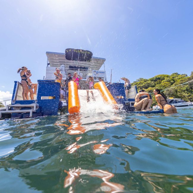 People enjoying water activities, some sliding down an inflatable slide from a boat into the water, with another boat and green trees in the background under a clear sky. Perfect for those looking to book a Sydney boat party hire or planning corporate boat events in Sydney.
