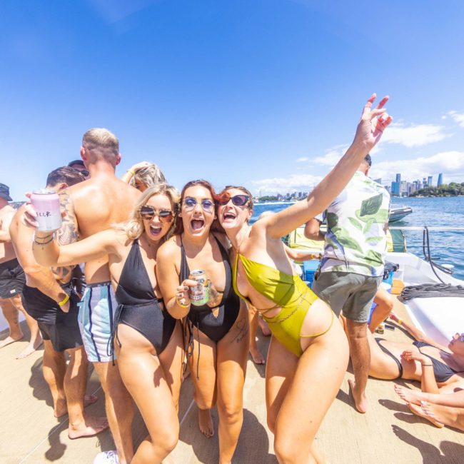 A group of people in swimwear enjoying a sunny day on a catamaran party in Sydney, with three women in the forefront smiling and holding drinks; cityscape visible in the background.