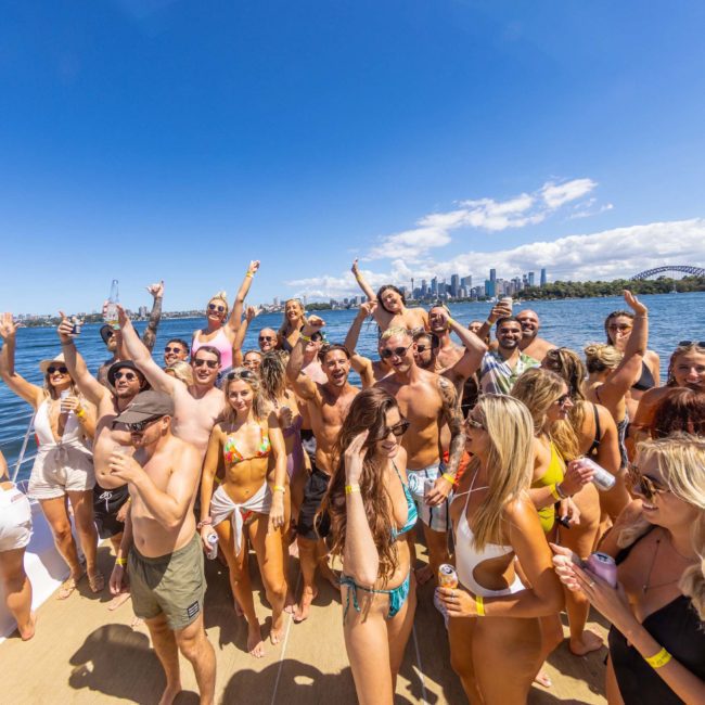 A group of people in swimwear are gathered on a luxury yacht under clear skies, with a city skyline and body of water in the background. Many are smiling, raising their arms, and appearing to celebrate.