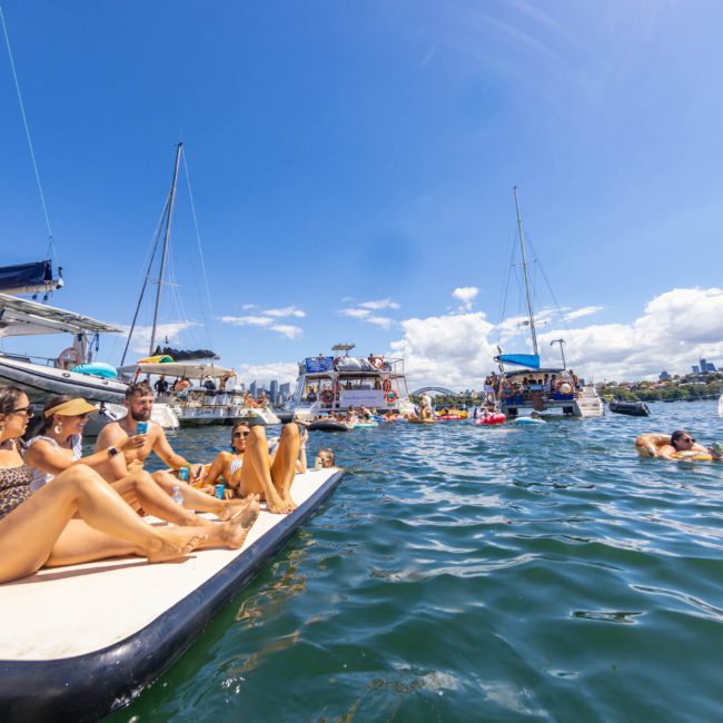 People relaxing on a floating mat near boats in a sunny harbor, some wearing swimsuits. In the background, other boats and swimmers enjoy the water during a vibrant catamaran party Sydney.