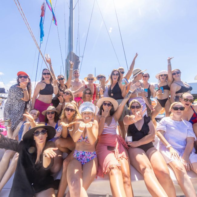 A large group of women pose together on a luxury yacht hire in Sydney under a sunny sky, many wearing hats and sunglasses.
