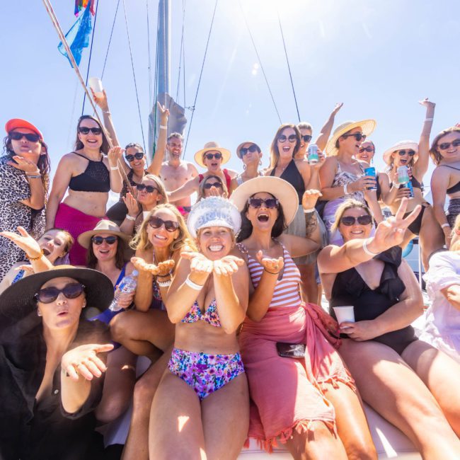 A group of people on a luxury yacht hire in Sydney posing for a photo, wearing summer attire including sunglasses, hats, and swimwear. Many are smiling and making cheerful gestures under a sunny sky.