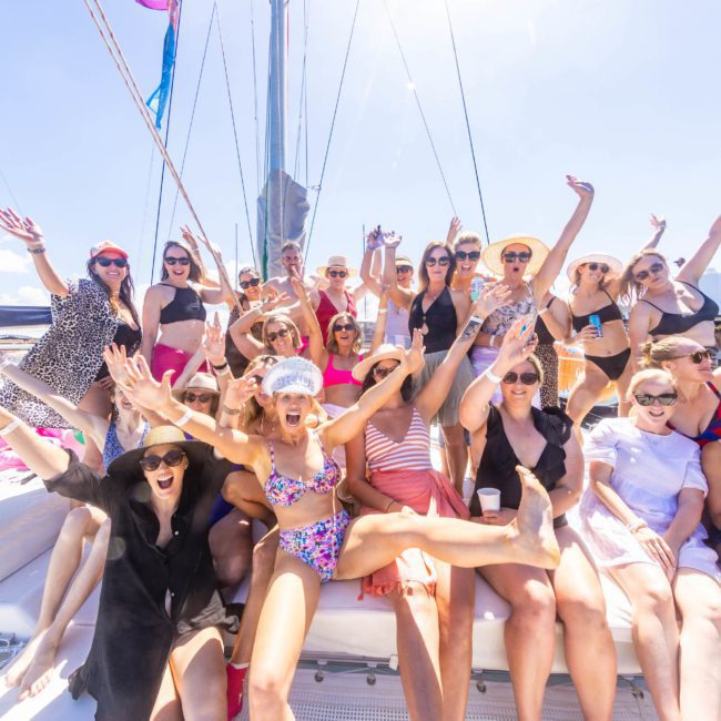 A large group of people in swimwear and hats cheerfully pose on a luxury yacht hire Sydney under clear skies.
