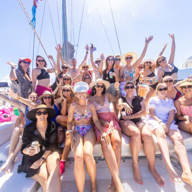 A group of people in swimwear and summer clothing pose together on a luxury yacht hire Sydney, raising their hands and smiling under a sunny sky.
