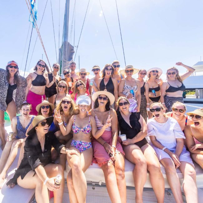 A group of people in swimwear and casual summer clothes pose for a photo on a boat under sunny skies, enjoying one of the best Sydney boat party hire experiences.