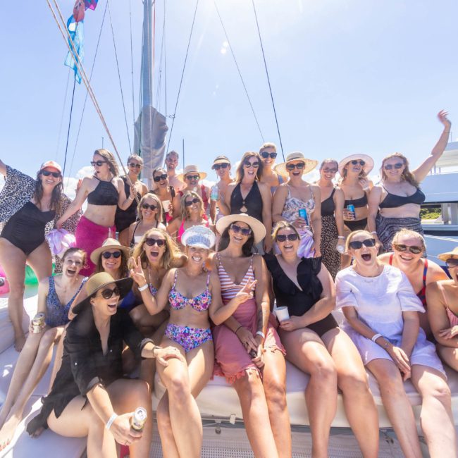 A large group of people on a private yacht charter in Sydney Harbour, smiling and posing for a picture under sunny skies. Some wear swimwear and hats, while others hold drinks. A watercraft is visible in the background.