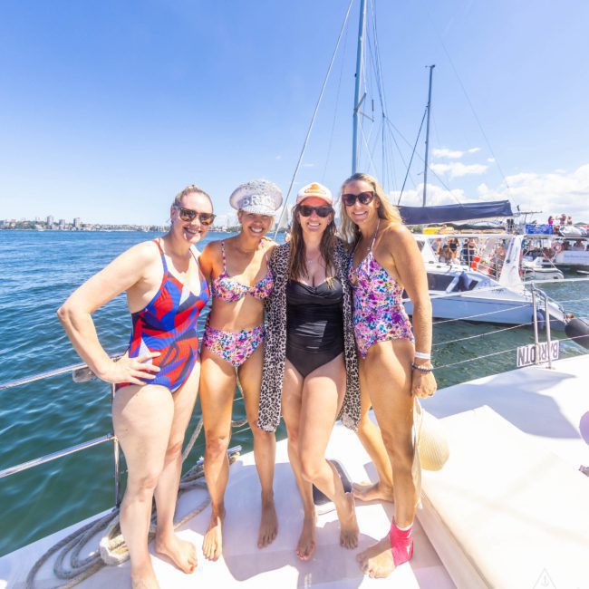 Four women in swimsuits stand on a boat deck with a marina and city skyline in the background, enjoying an amazing catamaran party in Sydney.