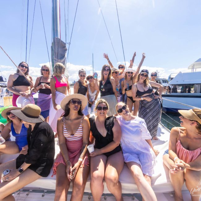 A group of women, dressed in summer outfits and hats, are gathered on a boat, smiling and posing for a photo with a marina and boats in the background during a catamaran party on Sydney Harbour.