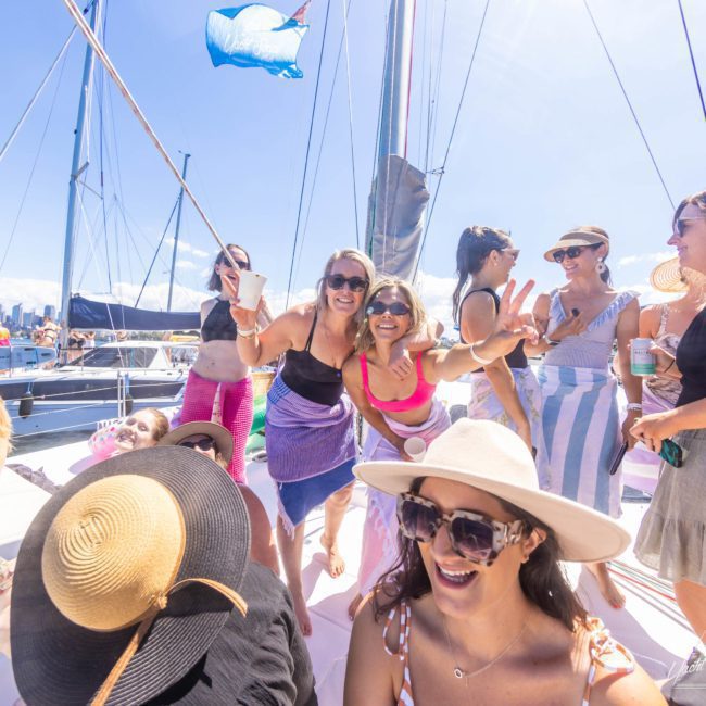 A group of people wearing summer clothing are on a sailboat, smiling and enjoying a sunny day. Some are holding drinks, with the backdrop featuring other boats and a bright blue sky—perfect for a private yacht charter Sydney Harbour or any Sydney boat party hire.