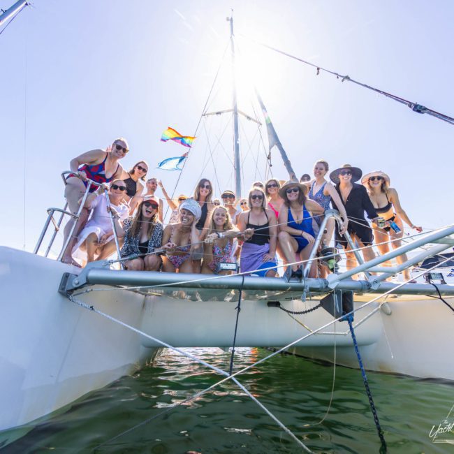 A group of people posing together at the front of a docked luxury yacht hire Sydney on a sunny day, with a clear blue sky and some colorful flags in the background.