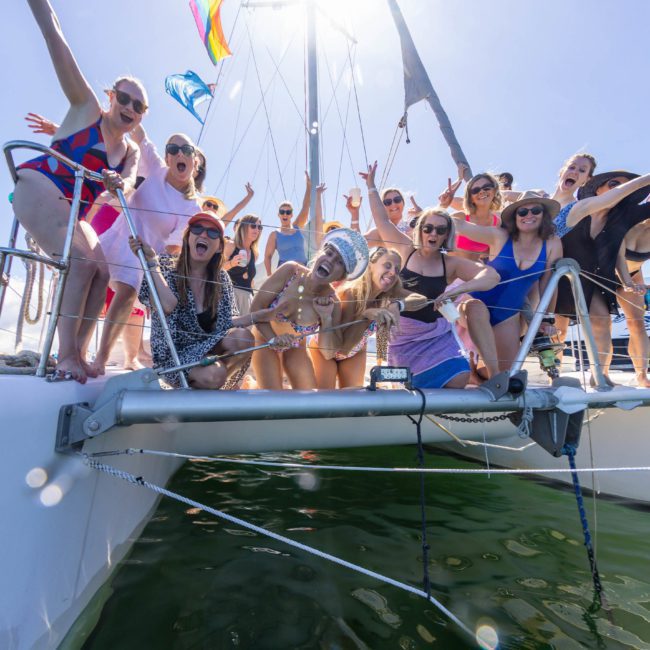 A group of people in swimwear are gathered at the stern of a private yacht charter on Sydney Harbour, smiling and posing for the camera on a sunny day.