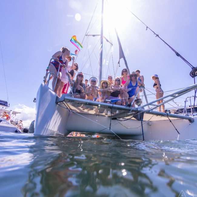 A group of people is gathered on a sailboat in sunny weather, some holding colorful flags. The boat is on calm water with more boats visible in the background—a perfect scene for a private yacht charter Sydney Harbour or a DJ boat hire Sydney celebration.