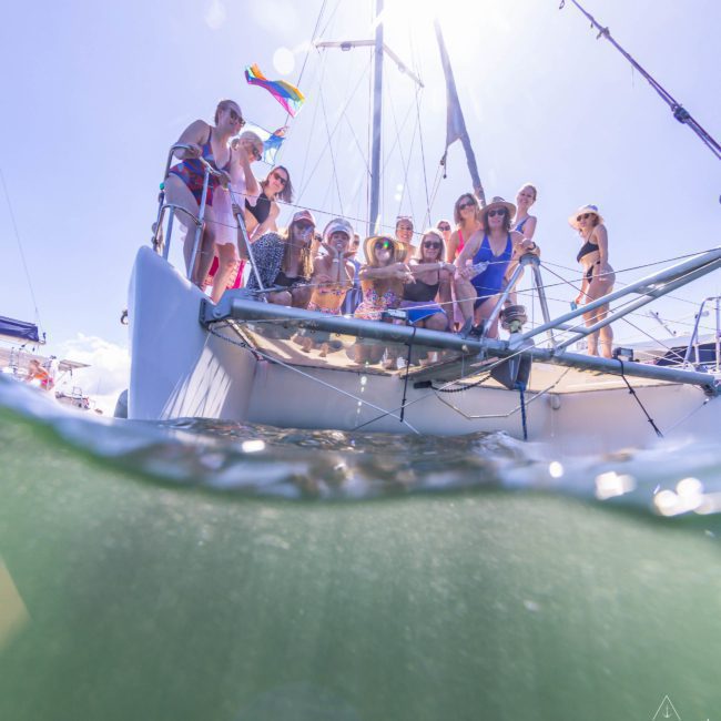 A group of people on a sailboat pose for a photo. Some are sitting and some are standing, with a rainbow flag visible in the background. The photo, taken partially from underwater, could be from a Sydney boat party hire event.