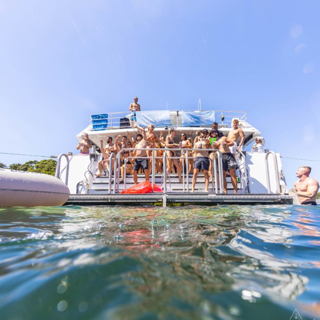 A group of people stand on a boat, some looking towards the water and others interacting. The boat is anchored, and the water is calm. A person is visible swimming near the boat during a lively catamaran party Sydney event.