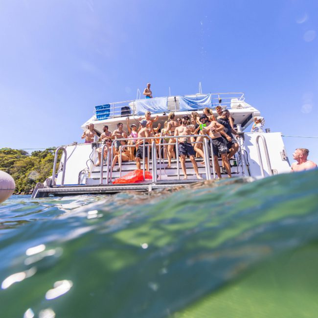 A group of people in swimsuits are standing on the back deck of a luxury yacht anchored in clear water on a sunny day. Some individuals are jumping into the water, enjoying the Sydney boat party hire experience.