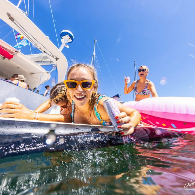 People enjoy a sunny day on a private yacht charter in Sydney Harbour, with one person in yellow sunglasses smiling at the camera and holding a can, while others in swimsuits relax on an inflatable float.