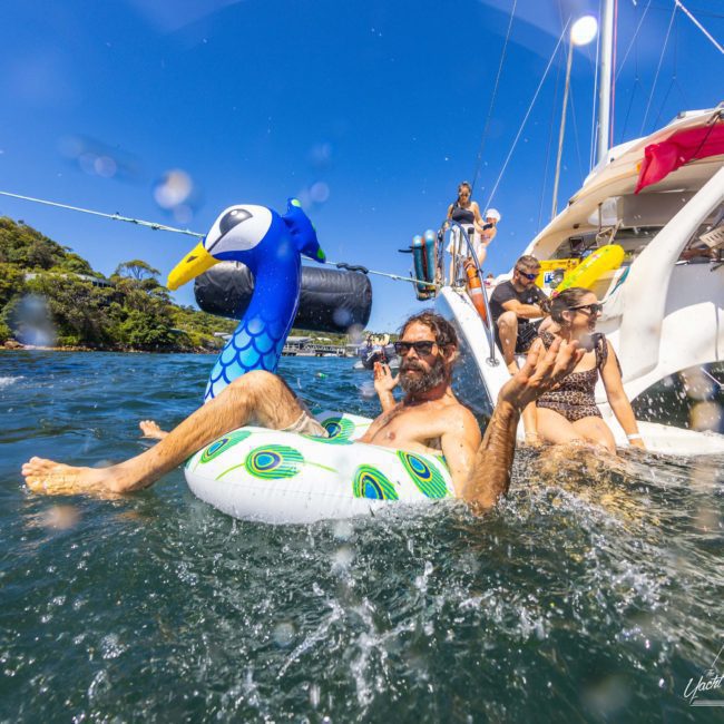 A man relaxes on a peacock-shaped inflatable tube in the water near a luxury yacht, with other people enjoying the sunny day on the boat.