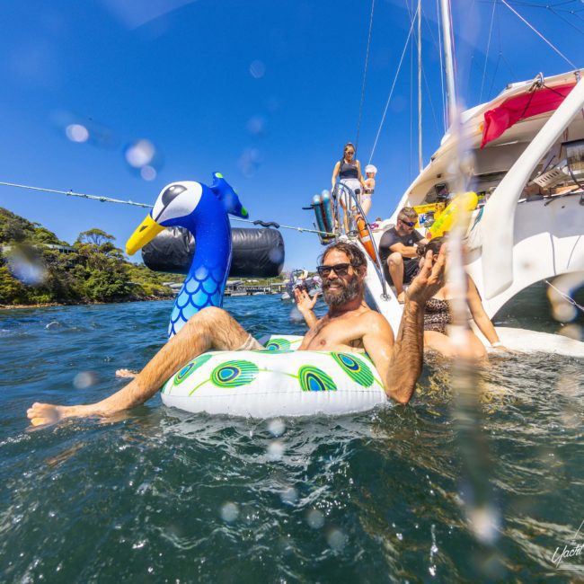 A group of people enjoys a sunny day on a private yacht charter in Sydney Harbour, with one man relaxing in the water on an inflatable peacock ring.