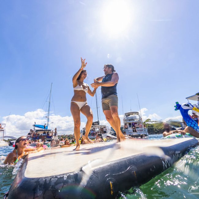 Two people dance on a floating platform in the water, surrounded by others swimming and enjoying a sunny day near boats, perfect for those interested in a luxury yacht hire in Sydney.
