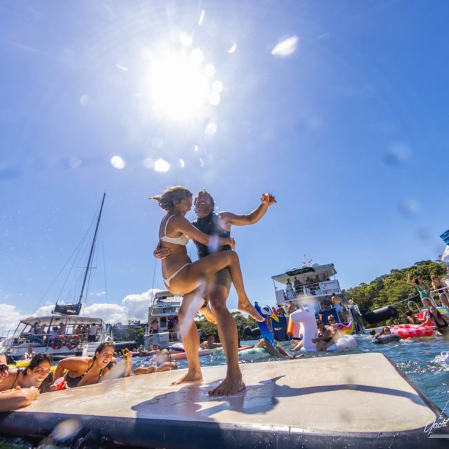 Two people are dancing on a floating platform in the water under a bright sun, surrounded by boats from a private yacht charter in Sydney Harbour and other people enjoying the water.