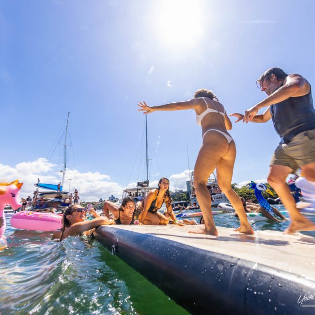 People are enjoying a sunny day on the water, with some standing on a platform and others in the water. A large inflatable flamingo is also visible. Sailboats, available for private yacht charter in Sydney Harbour, are anchored in the background.