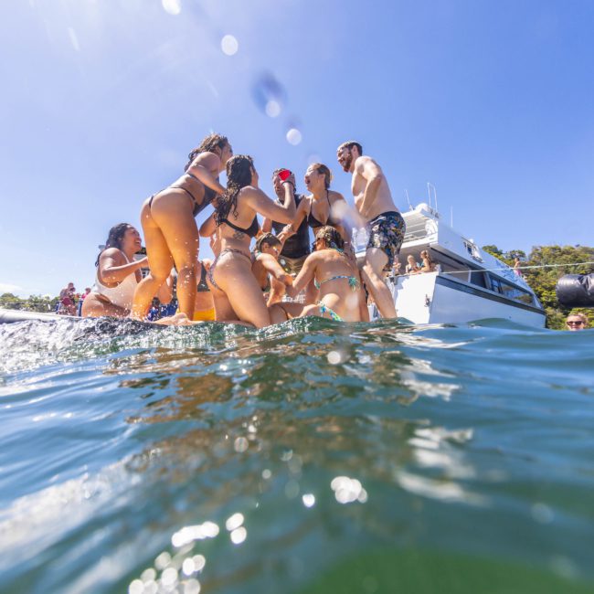 A group of people in swimwear are gathered on a floating platform in the water near a yacht on a sunny day, enjoying a lively Sydney boat party hire. Some are standing and talking, while others are partially submerged in the water.