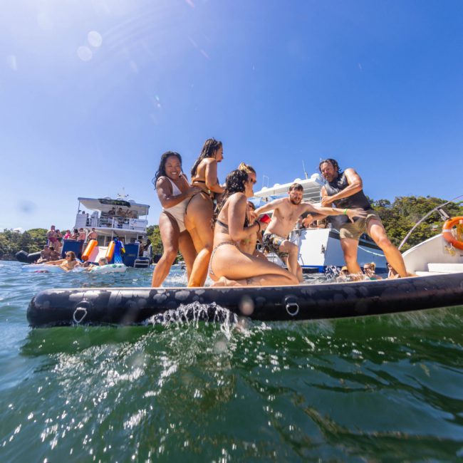 A group of people in swimwear are having fun on an inflatable platform in the water near a luxury yacht hire Sydney. Several others are visible on the yacht in the background.