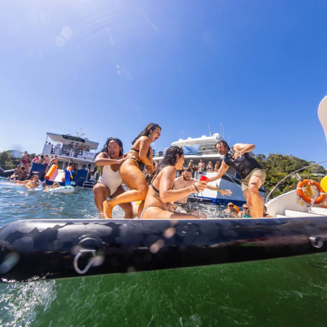 A group of people in swimwear are playing and balancing on an inflatable raft in the water near boats on a sunny day, enjoying their private yacht charter Sydney Harbour.