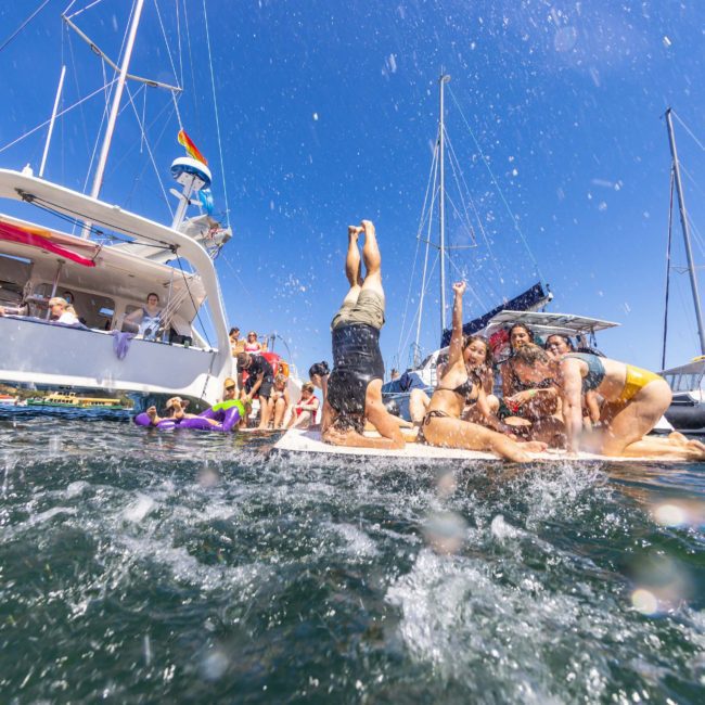 A group of people enjoys a sunny day on the water, playing and splashing near sailboats. Some individuals kneel and cheer while others swim or relax on the boat during a luxurious catamaran party Sydney experience.