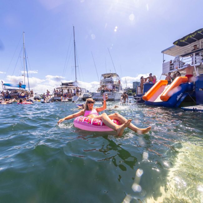 Person floats on an inflatable ring in the water near several anchored boats with people, enjoying a sunny day under a blue sky. An inflatable slide is visible on one boat, perfect for a fun Sydney boat party hire.