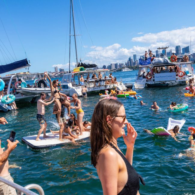 A group of people enjoy a sunny day on the water, surrounded by boats and inflatables. Some are swimming, while others relax on rafts. The skyline of a city can be seen in the background, emphasizing the allure of a luxury yacht hire Sydney has to offer for an unforgettable experience.
