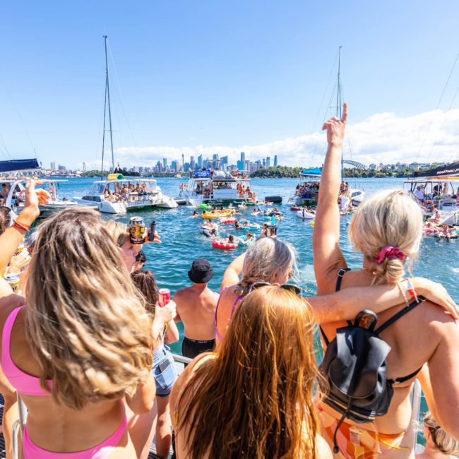 A large group of people in swimsuits enjoy a lively boat party on a sunny day, with several boats and people on inflatable floats in the water, and a cityscape visible in the background, showcasing the perfect setting for a catamaran party Sydney.