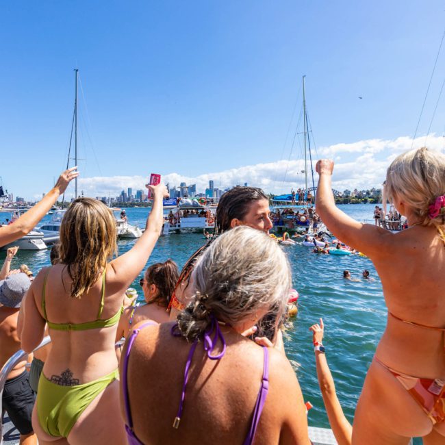 A group of people in swimwear stand on a dock and raise their hands, facing a body of water with boats and other swimmers. A city skyline is visible in the background under a clear blue sky, creating the perfect setting for corporate boat events Sydney and private yacht charters.