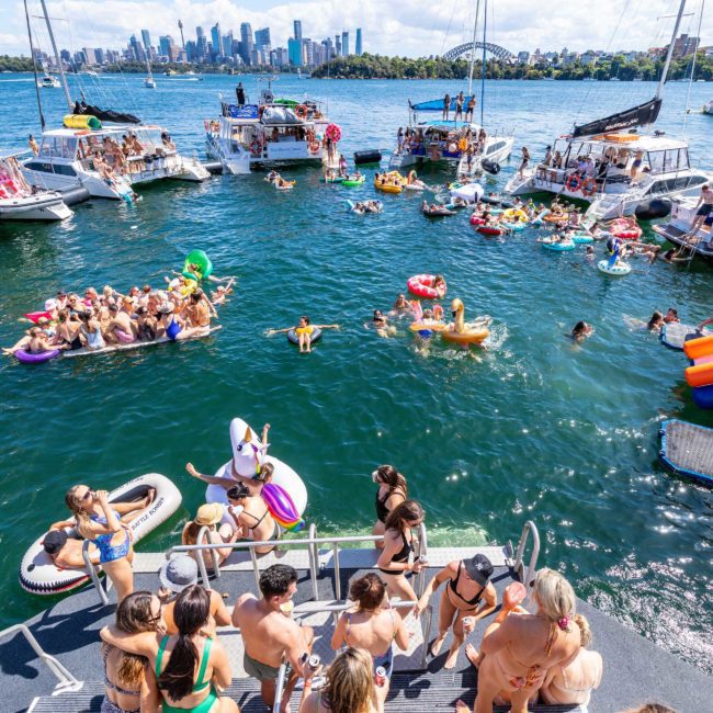 People aboard yachts and various inflatables are enjoying a sunny, festive gathering on the water, in front of a city skyline, highlighting the appeal of luxury yacht hire Sydney.