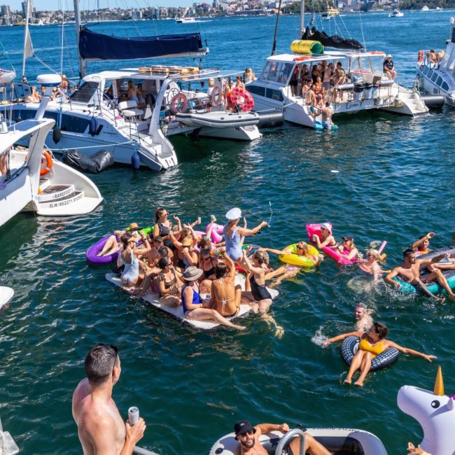 A group of people enjoying a sunny day on boats and inflatable floats in a body of water, with some swimming and socializing. Various boats, including private yacht charters in Sydney Harbour, are anchored nearby.