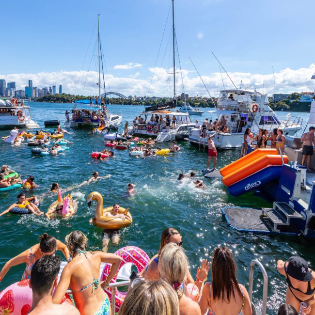 People in swimsuits enjoying a sunny day on the water with inflatables and boats anchored nearby. Some are swimming, while others relax on boats rented through Sydney boat party hire or use a floating water slide. City skyline in the background.
